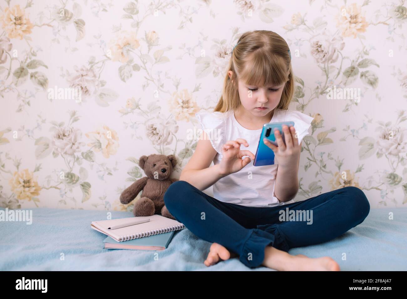 Une petite fille s'assoit sur le lit dans la chambre élégante, tenant le téléphone et lit quelque chose dans le smartphone. Concept de communication Banque D'Images