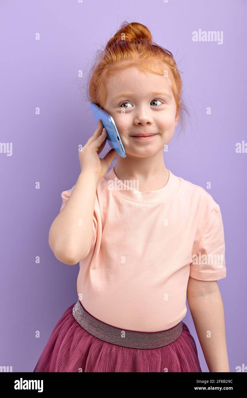 REDHEAD enfant fille communique sur téléphone mobile, sourit et parle isolé sur fond violet. Gros plan sourire enfant parlant sur smartphone. Le péché des gens Banque D'Images