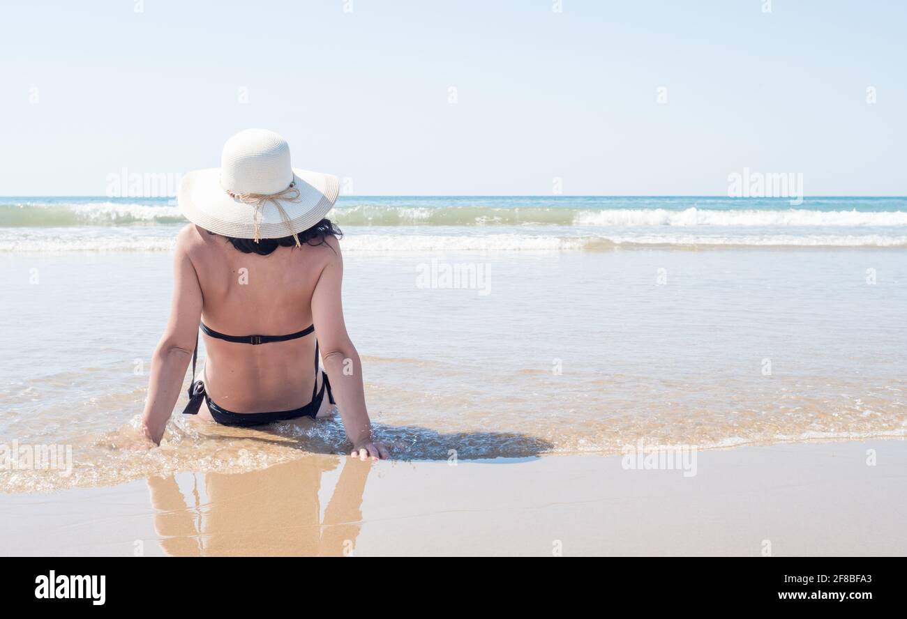une jeune femme en chapeau et en bikini noir se détend l'eau sur la plage en regardant l'horizon Banque D'Images
