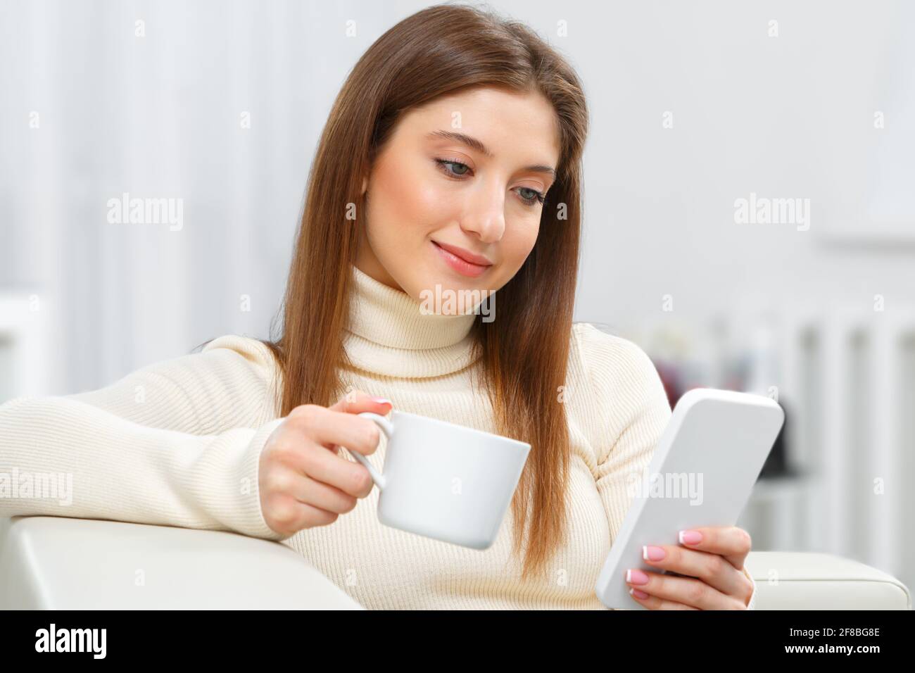 Belle petite fille est assise sur le canapé à la maison avec une tasse de café. Une jeune femme heureuse regarde dans l'écran du téléphone cellulaire. Banque D'Images