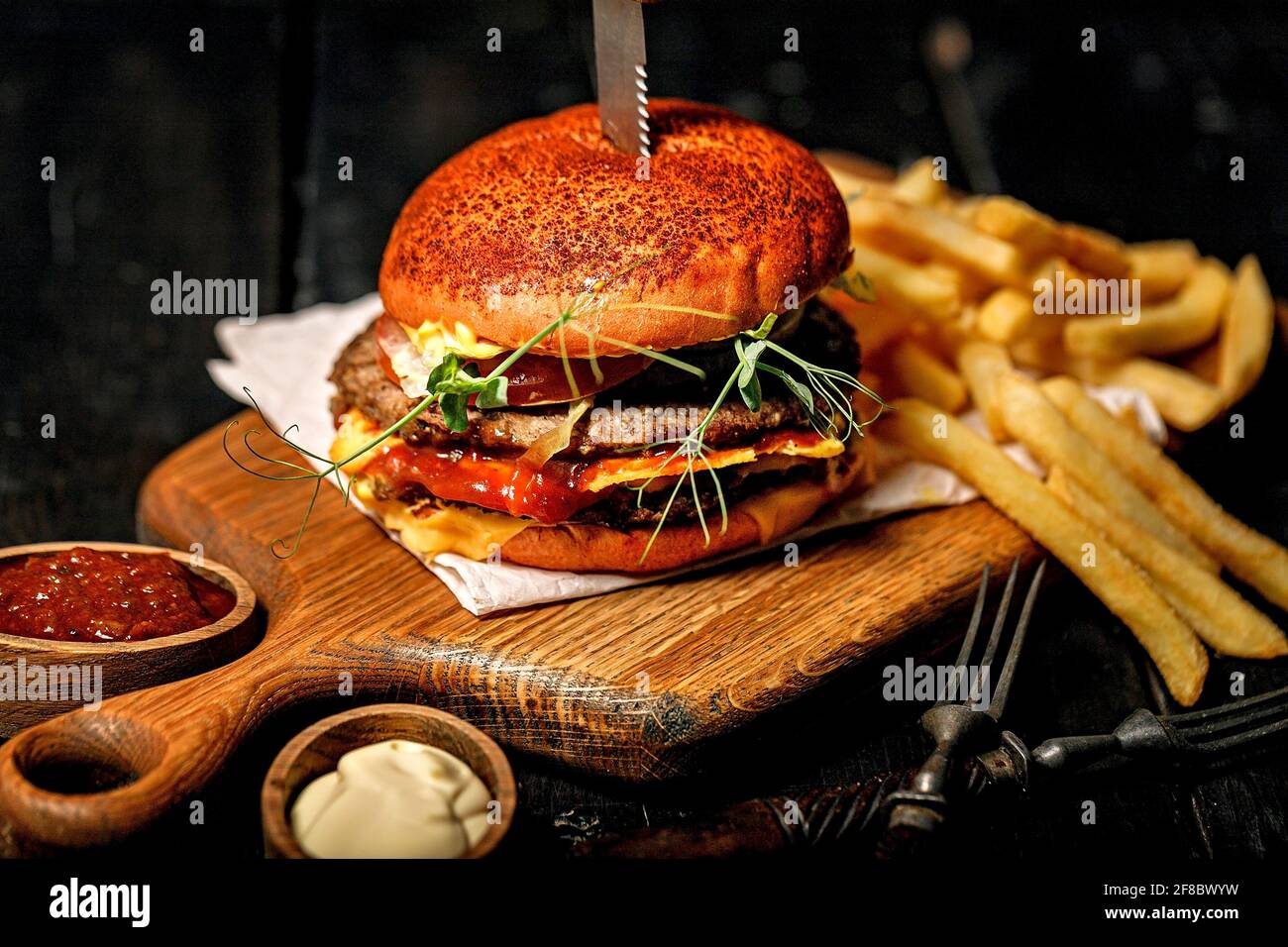Un hamburger de bœuf double appétissant et une portion de frites sur une planche à découper en bois. Arrière-plan rustique sombre. Photographie de la restauration rapide STILL Life. Gros plan Banque D'Images