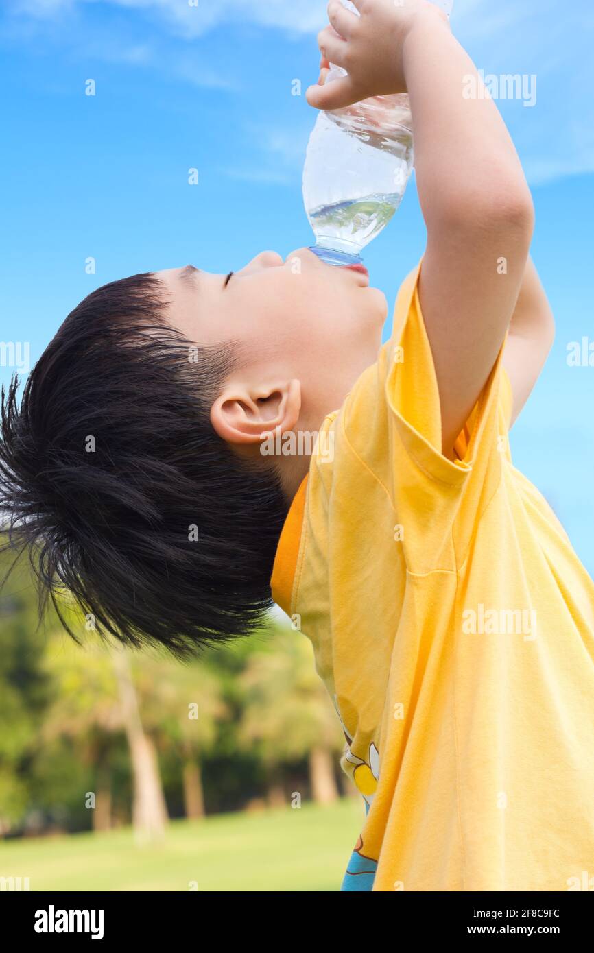 Un petit garçon asiatique boit de l'eau à partir d'une bouteille en plastique avec le soif, après avoir été fatigué d'une poule mouillée dans le parc, sous la lumière du soleil et le ciel bleu Backgrou Banque D'Images