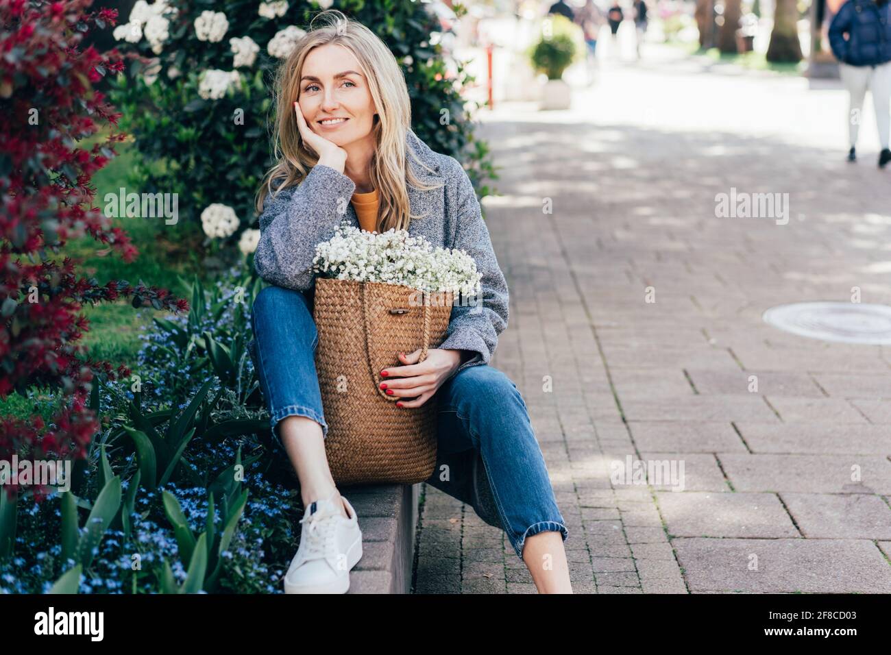 Jolie femme souriante, sous un manteau gris, assise près du Lit de fleur..femme caucasienne élégante posant à l'extérieur parmi les fleurs et sac de maintien avec Banque D'Images