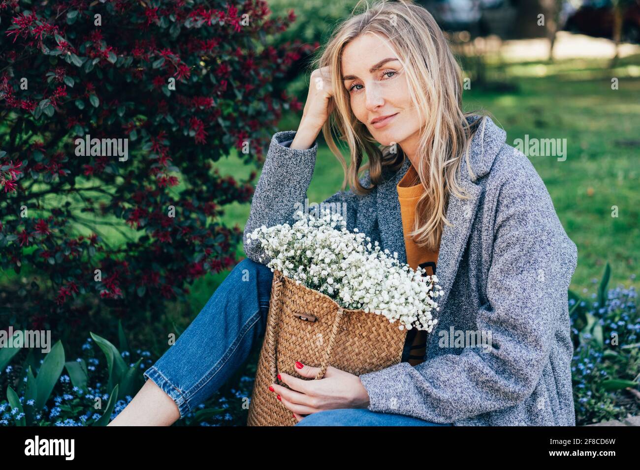 Charmante femme européenne à la mode avec un sac en osier et des fleurs de gitsophila. La fille est assise sur le parapet dans le parc. Portrait de printemps d'un hipster Banque D'Images