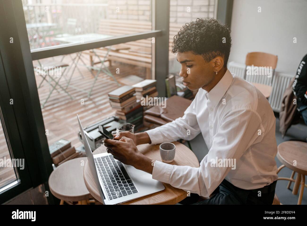 Un jeune homme noir est assis à la table avec un ordinateur portable, buvant du thé et utilisant un smartphone. Le gars dans le café. Travailler à distance en dehors du bureau Banque D'Images
