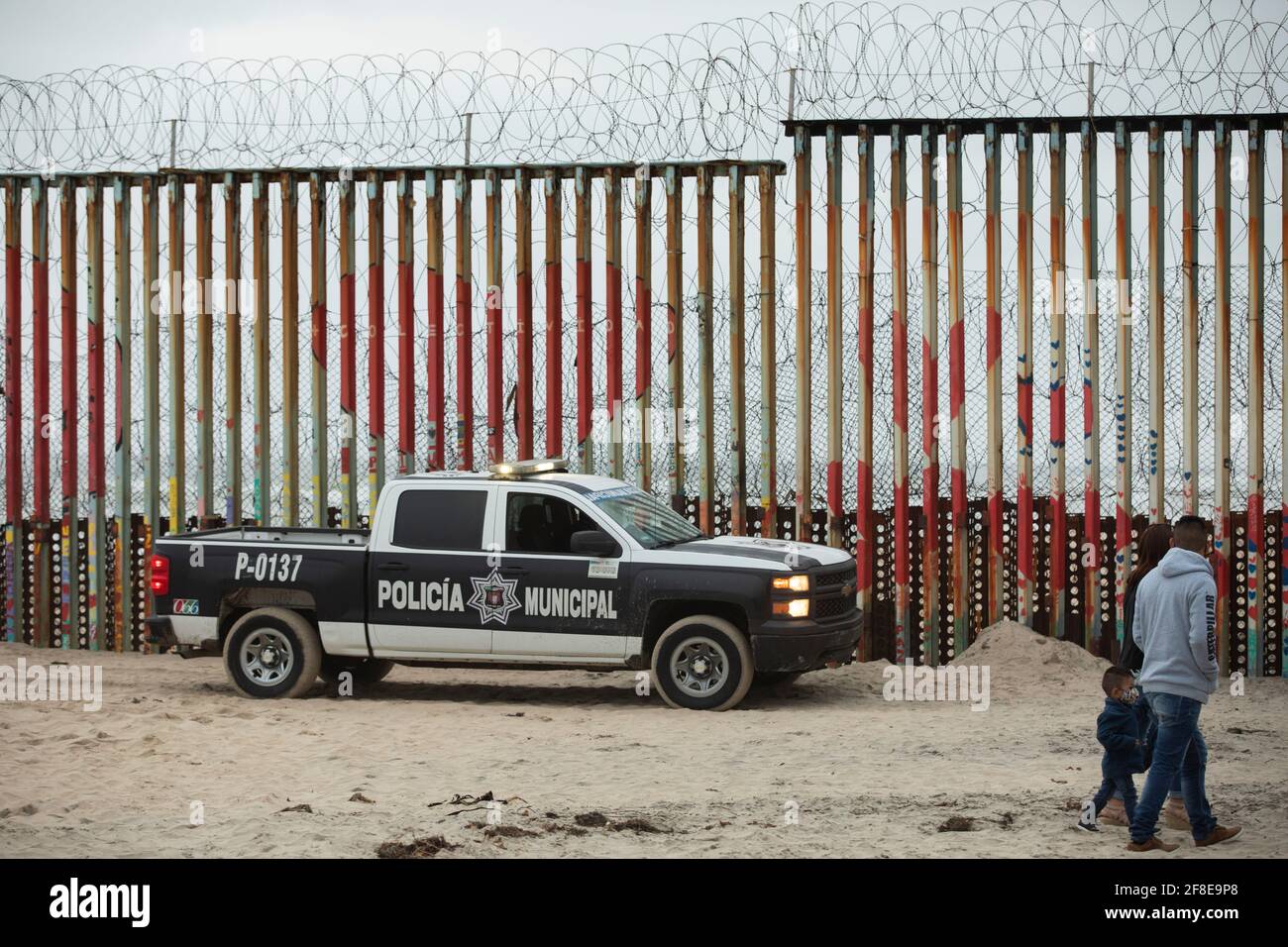 Tijuana, Basse-Californie, Mexique - le 11 avril 2021 : une famille marche devant le mur frontalier des États-Unis-Mexique tandis qu'une voiture de police municipale patrouille sur l'allongsid Banque D'Images