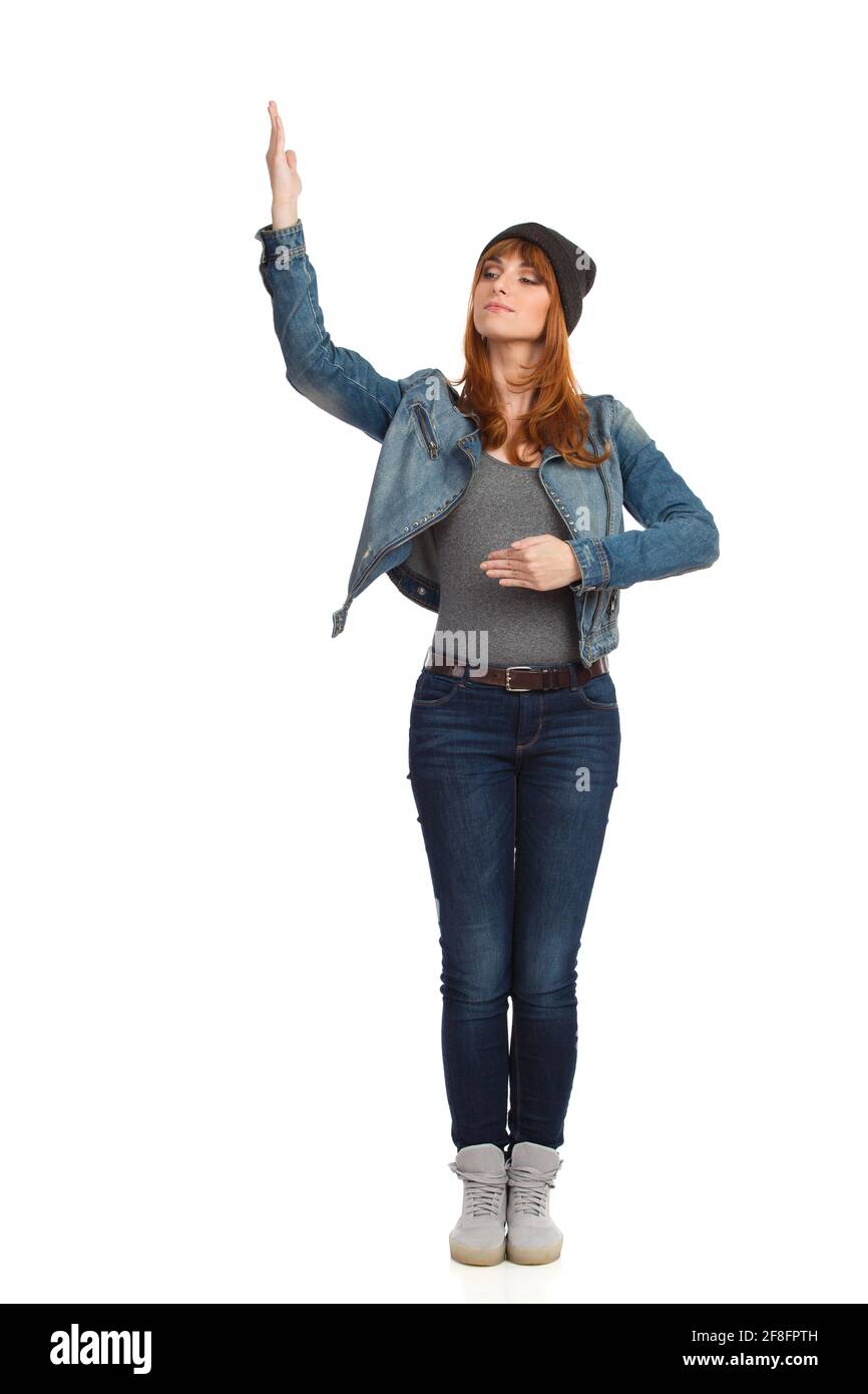 Jeune femme sérieuse en veste de Jean non boutonnée, est debout, tenant le bras relevé et regardant loin. Vue avant. Prise de vue en studio sur toute la longueur isolée sur le coup Banque D'Images