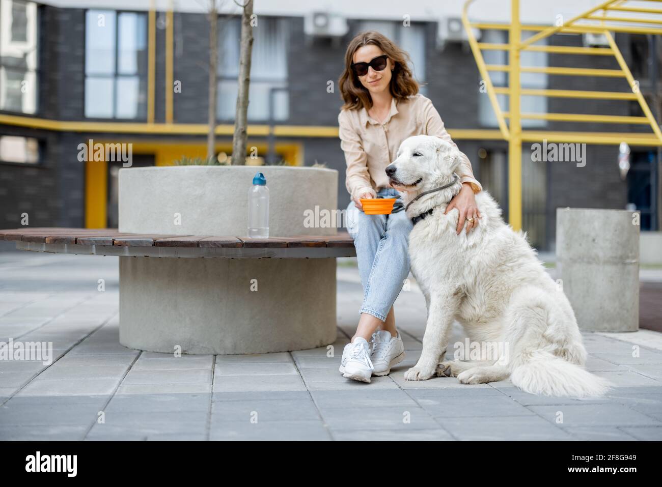 Une femme donne de l'eau dans un bol à son grand chien blanc tout en étant assise sur un banc dans la cour de la résidence. Soins pour animaux, amoureux des animaux. Banque D'Images