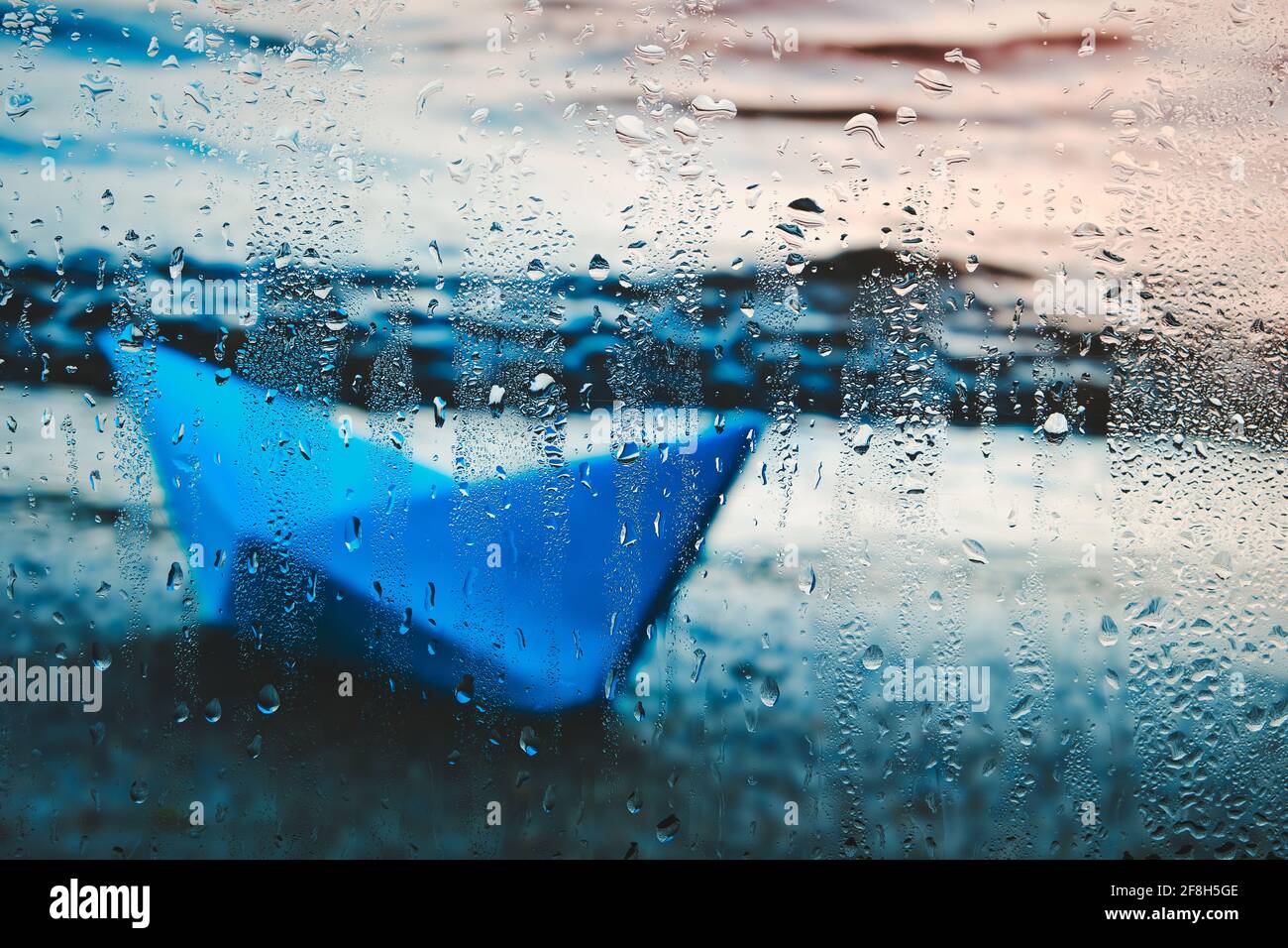 Le bateau bleu papier est lavé sur la plage au coucher du soleil, derrière le verre humide en gouttes de pluie Banque D'Images