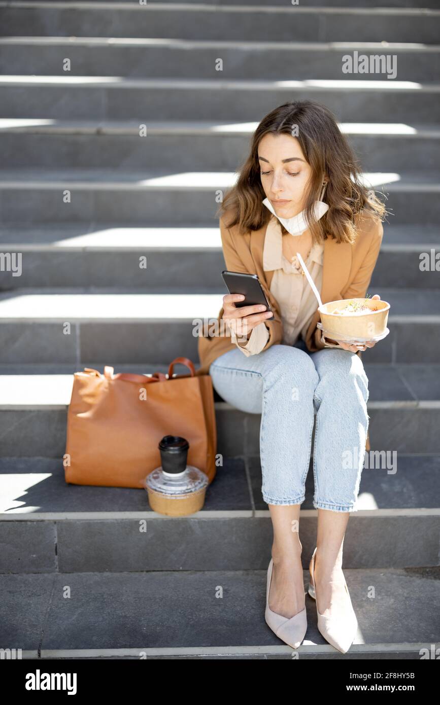 Jolie femme ont un déjeuner extérieur près du bâtiment de bureau tout en étant assise sur les escaliers et en utilisant le téléphone. Repas sain et café à emporter pour les travailleurs. Avec un masque de protection abaissé sur son menton Banque D'Images