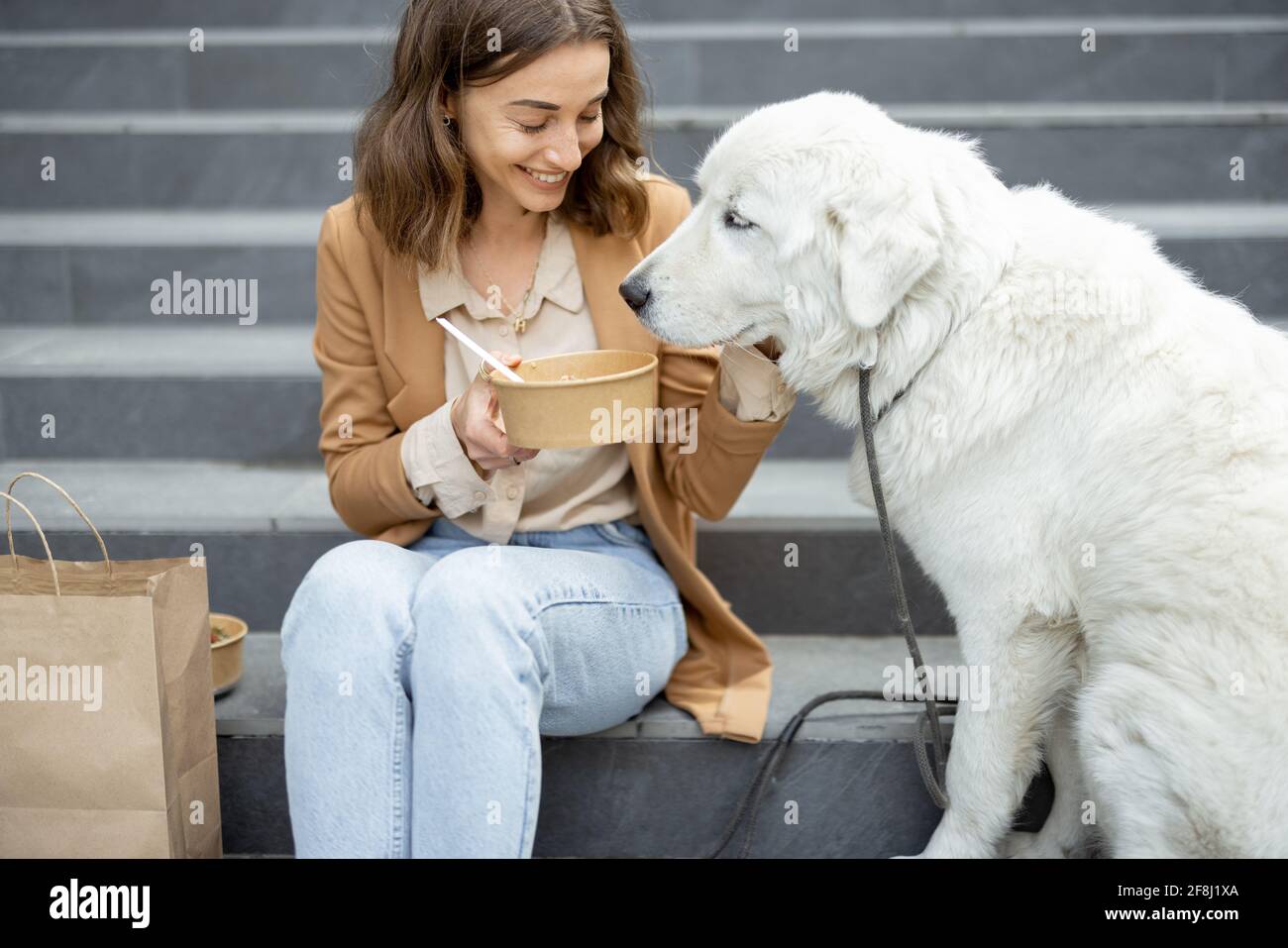 Jolie femme a déjeuner à l'extérieur près du bâtiment de bureau avec son grand chien blanc tout en étant assis sur les escaliers. Partage la nourriture avec un chien. Animaux acceptés et concept de soins pour animaux. Amoureux des animaux. Banque D'Images