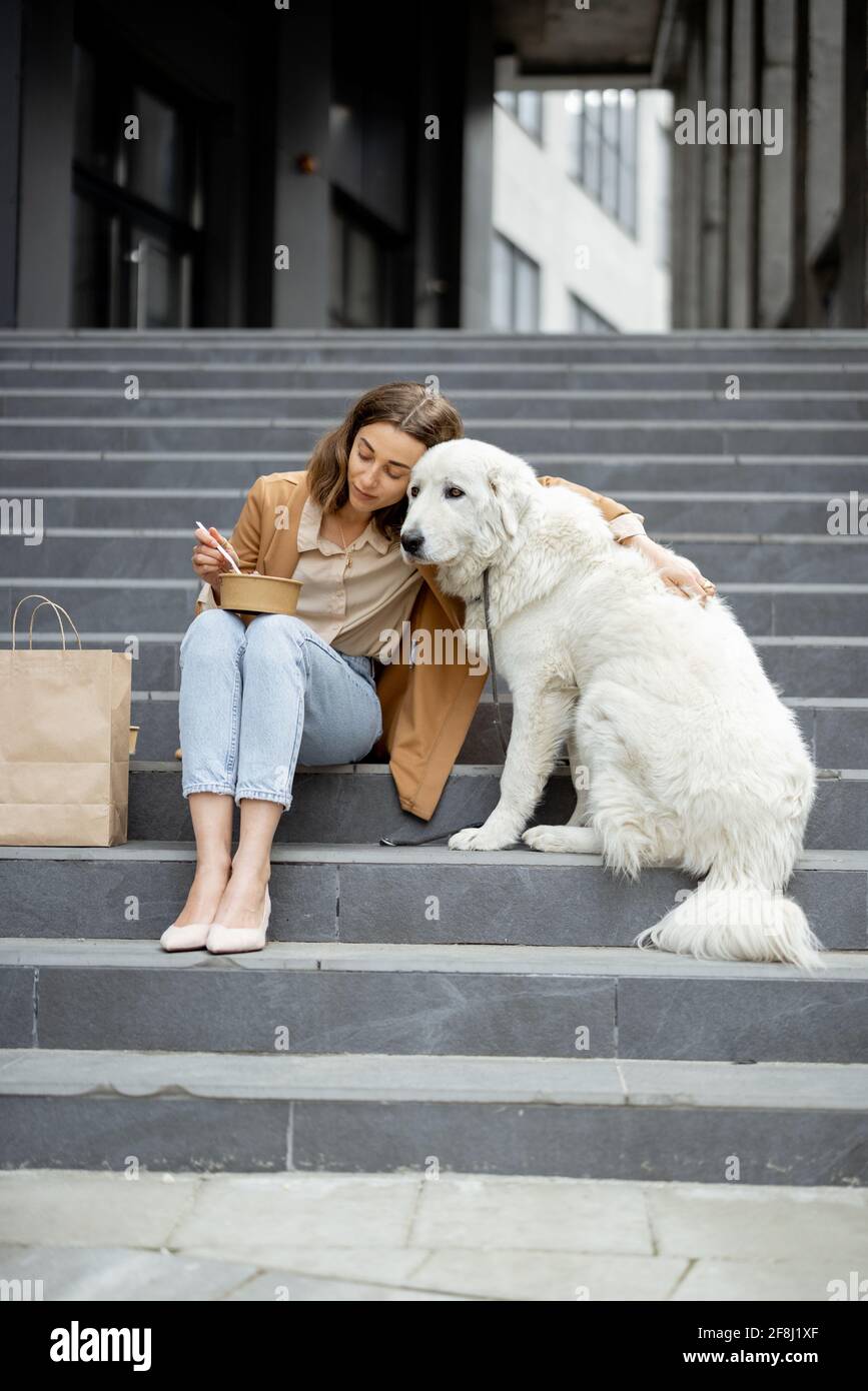 Jolie femme a un déjeuner à l'extérieur près du bâtiment de bureau avec son grand chien blanc tout en étant assis sur les escaliers, elle enchoit son chien. Animaux acceptés et concept de soins pour animaux. Amoureux des animaux. Banque D'Images