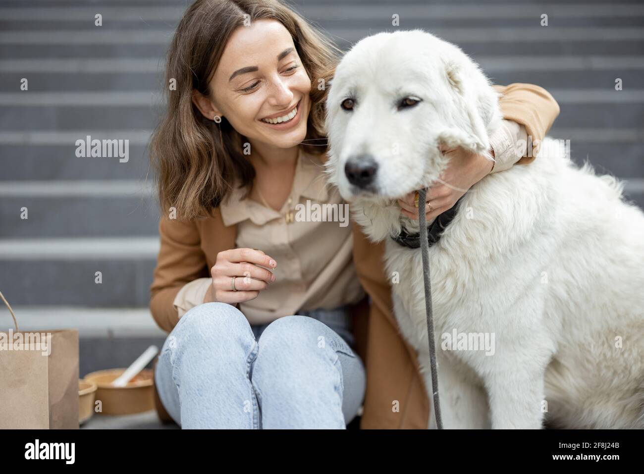 Jolie femme a un déjeuner à l'extérieur près du bâtiment de bureau avec son grand chien blanc tout en étant assis sur les escaliers, elle enchoit son chien. Animaux acceptés et concept de soins pour animaux. Amoureux des animaux. Banque D'Images