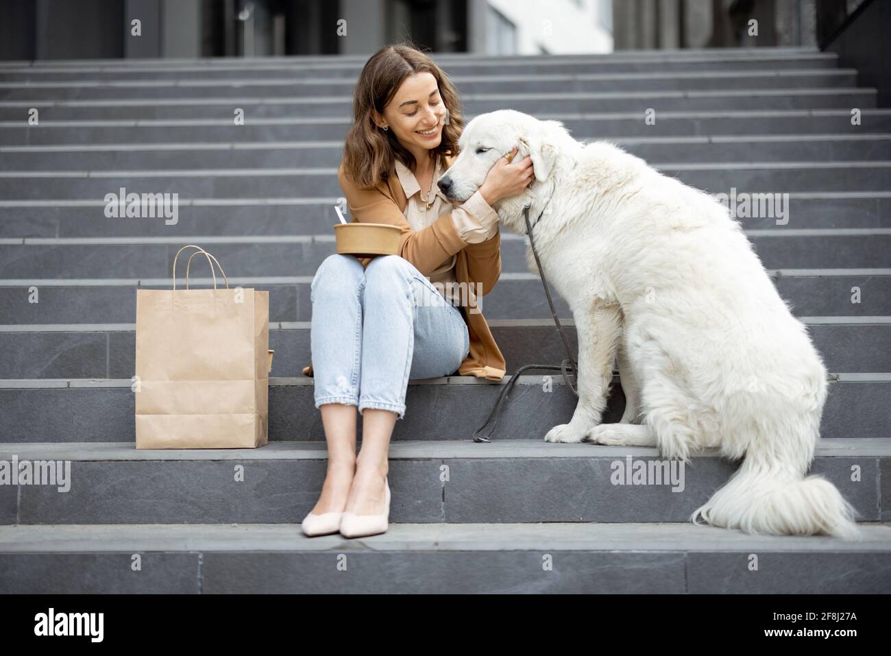 Jolie femme a déjeuner à l'extérieur près du bâtiment de bureau avec son grand chien blanc tout en étant assis sur les escaliers. Animaux acceptés et concept de soins pour animaux. Amoureux des animaux. Banque D'Images