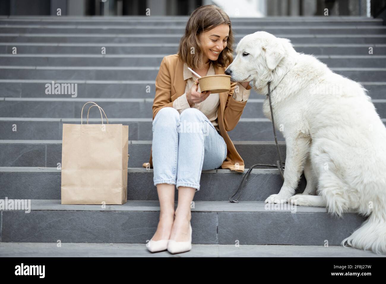Jolie femme a déjeuner à l'extérieur près du bâtiment de bureau avec son grand chien blanc tout en étant assis sur les escaliers. Partage la nourriture avec un chien. Animaux acceptés et concept de soins pour animaux. Amoureux des animaux. Banque D'Images