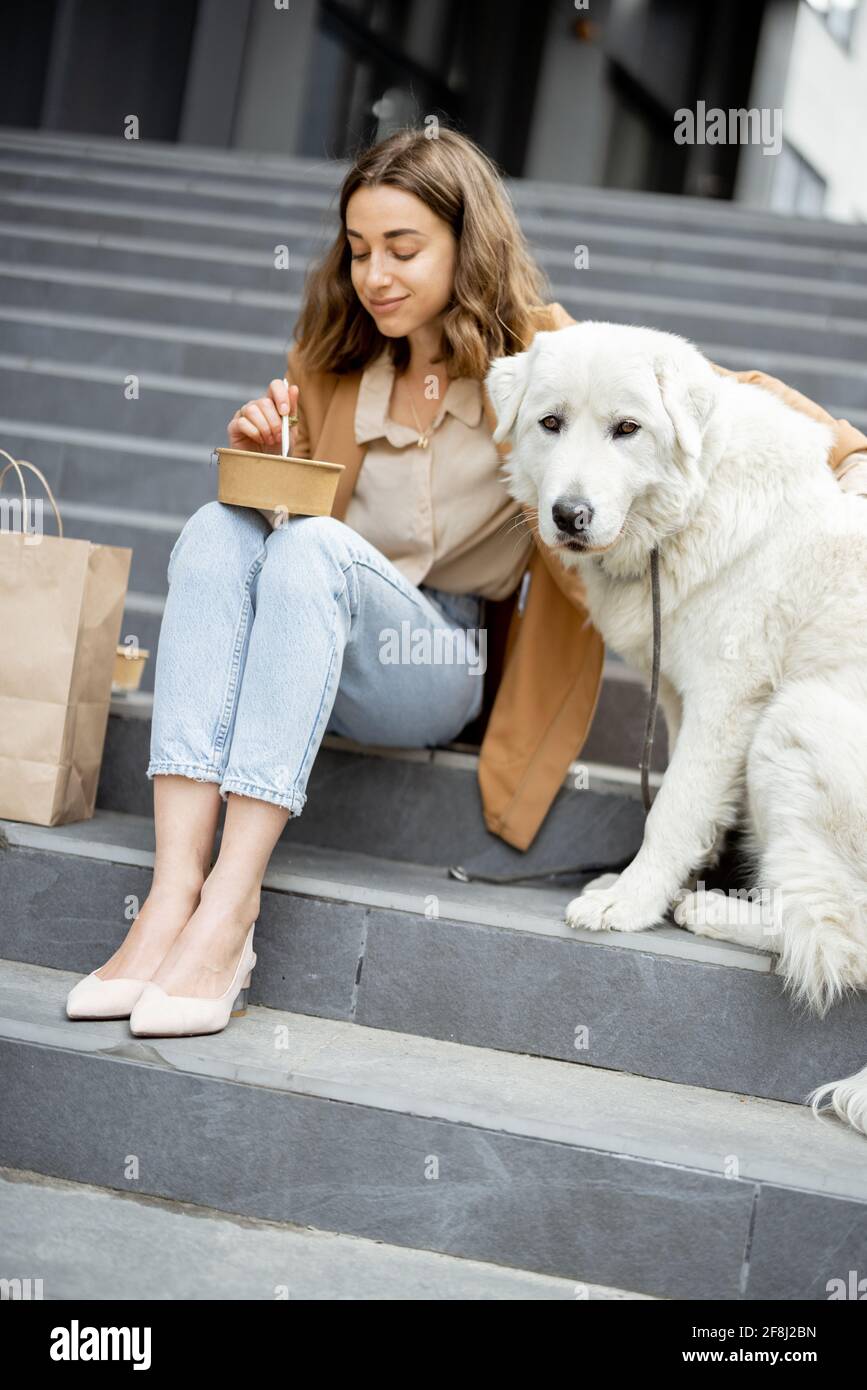 Jolie femme a déjeuner à l'extérieur près du bâtiment de bureau avec son grand chien blanc tout en étant assis sur les escaliers. Animaux acceptés et concept de soins pour animaux. Amoureux des animaux. Banque D'Images
