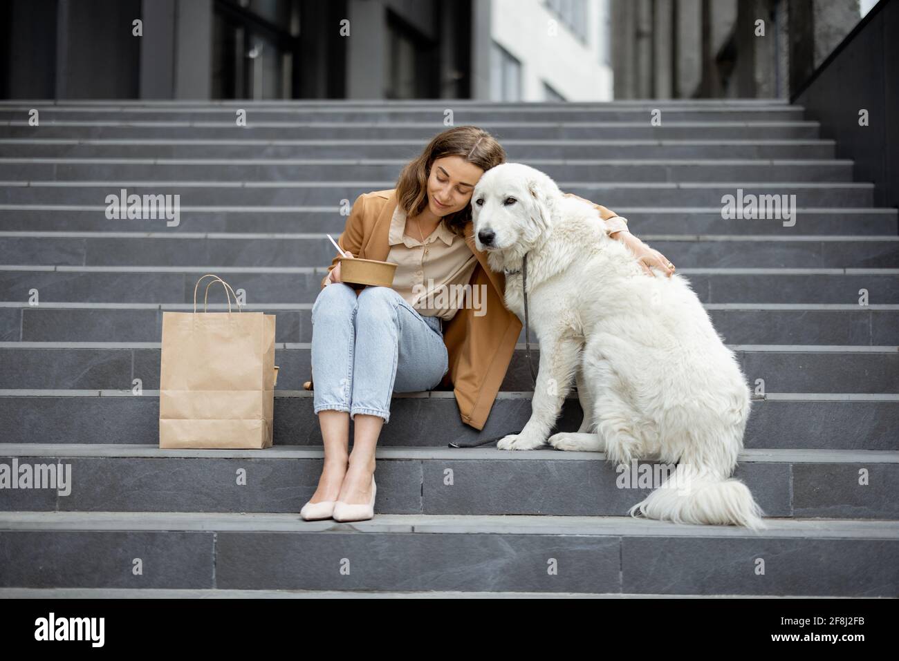 Jolie femme a un déjeuner à l'extérieur près du bâtiment de bureau avec son grand chien blanc tout en étant assis sur les escaliers, elle enchoit son chien. Animaux acceptés et concept de soins pour animaux. Amoureux des animaux. Banque D'Images