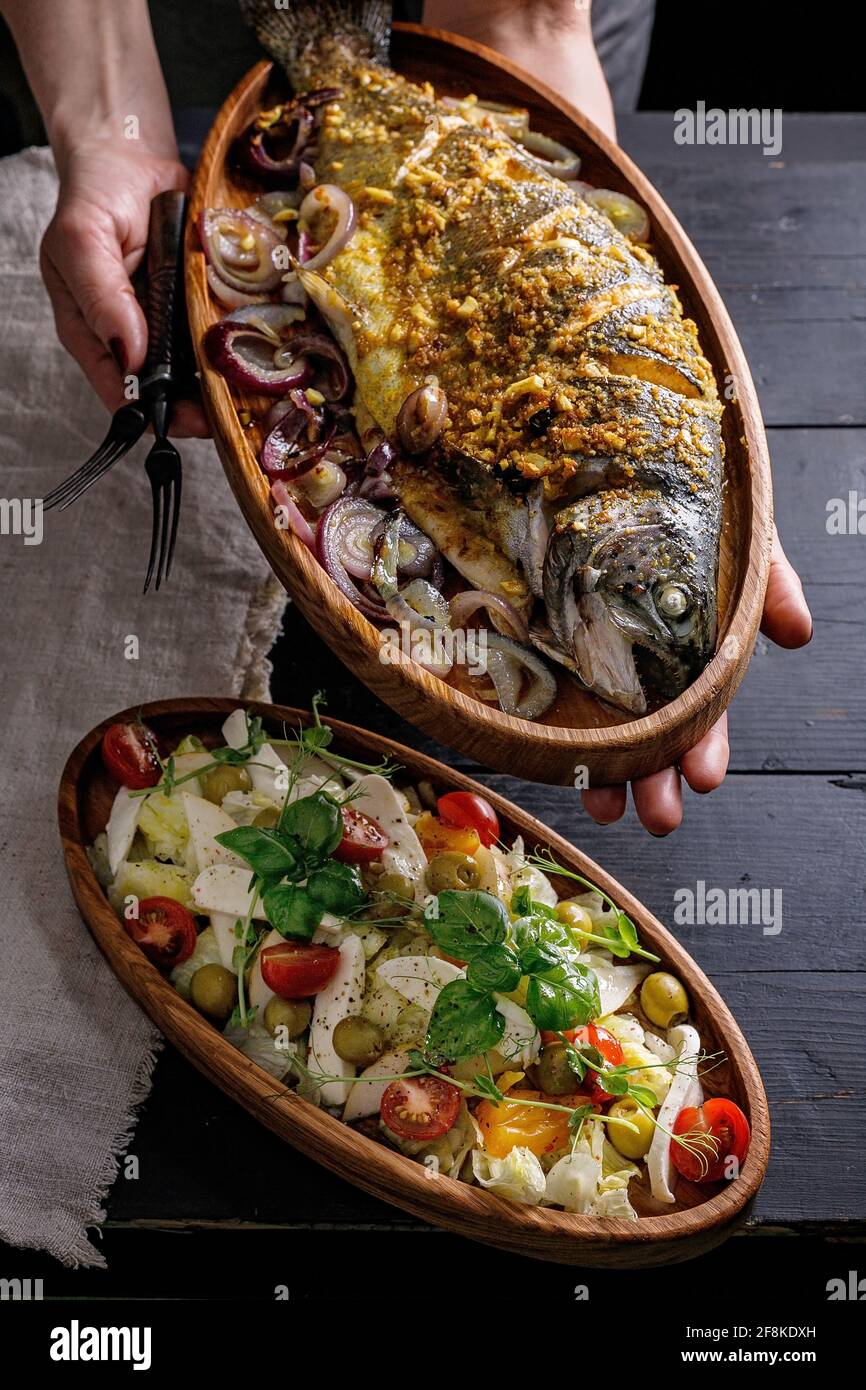 Les mains des femmes tiennent un plat en bois avec du poisson grillé. Salade de fruits de mer et de légumes. Un déjeuner délicieux et sain. Régime méditerranéen. Tir vertical. Nourriture p Banque D'Images