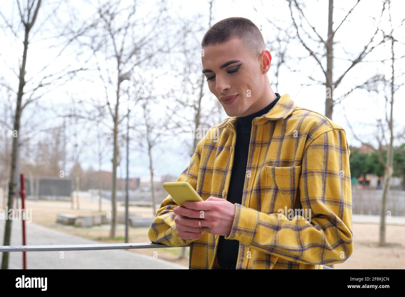 Jeune personne non binaire vérifiant son smartphone et souriant. Faites le maquillage de la personne androgyne. Banque D'Images