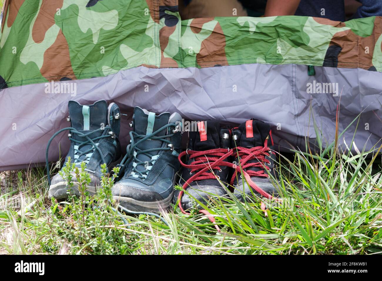 Deux paires de bottes de randonnée ou de marche pour enfants laissées bien alignées à l'extérieur d'une tente de camping camouflée sur un terrain d'herbe. Banque D'Images