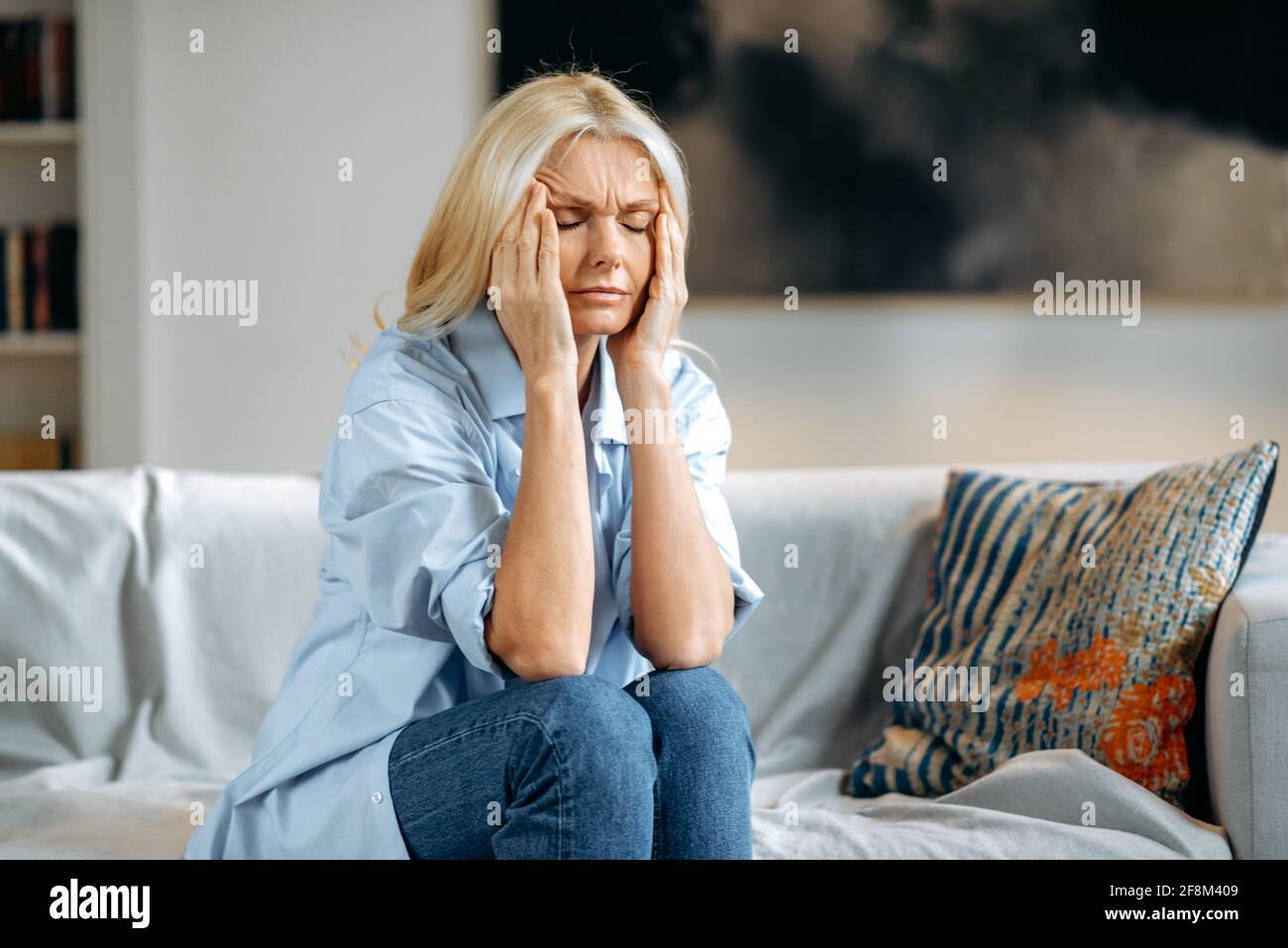 Une femme blanche blonde d'âge moyen est assise sur un canapé dans le salon  en tenant la tête avec ses mains, se sent malheureuse en raison de maux de  tête, de troubles