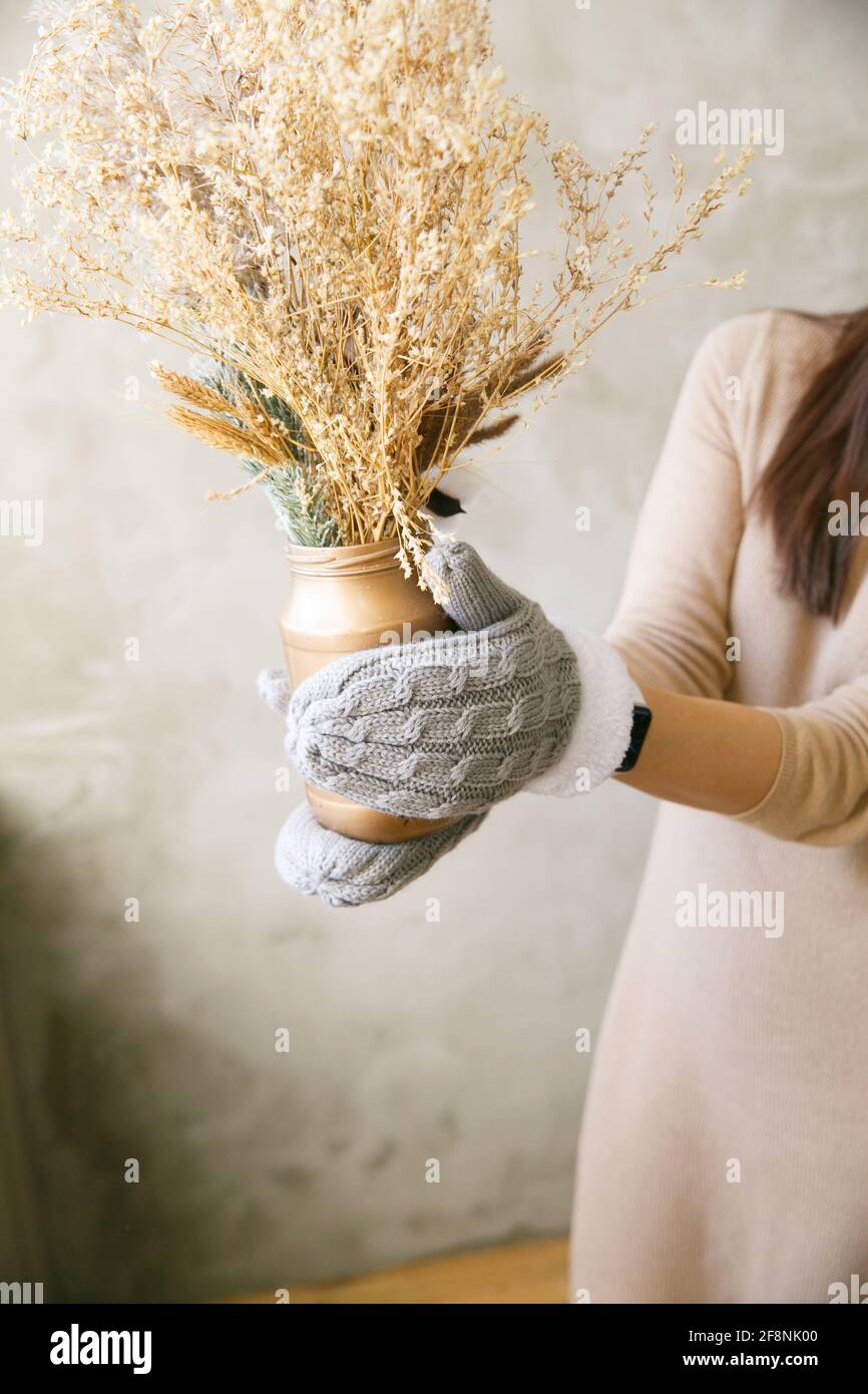 Une fille en moufles tient un vase avec des fleurs séchées près de la fenêtre le matin Banque D'Images