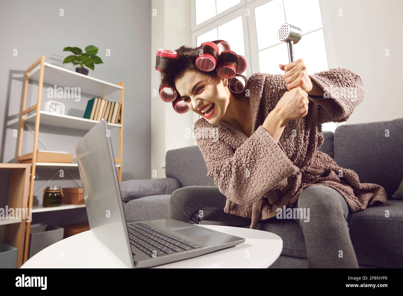 Furieuse femme bouleversée dans les rouleaux de cheveux détruisant son ordinateur portable avec un marteau Banque D'Images