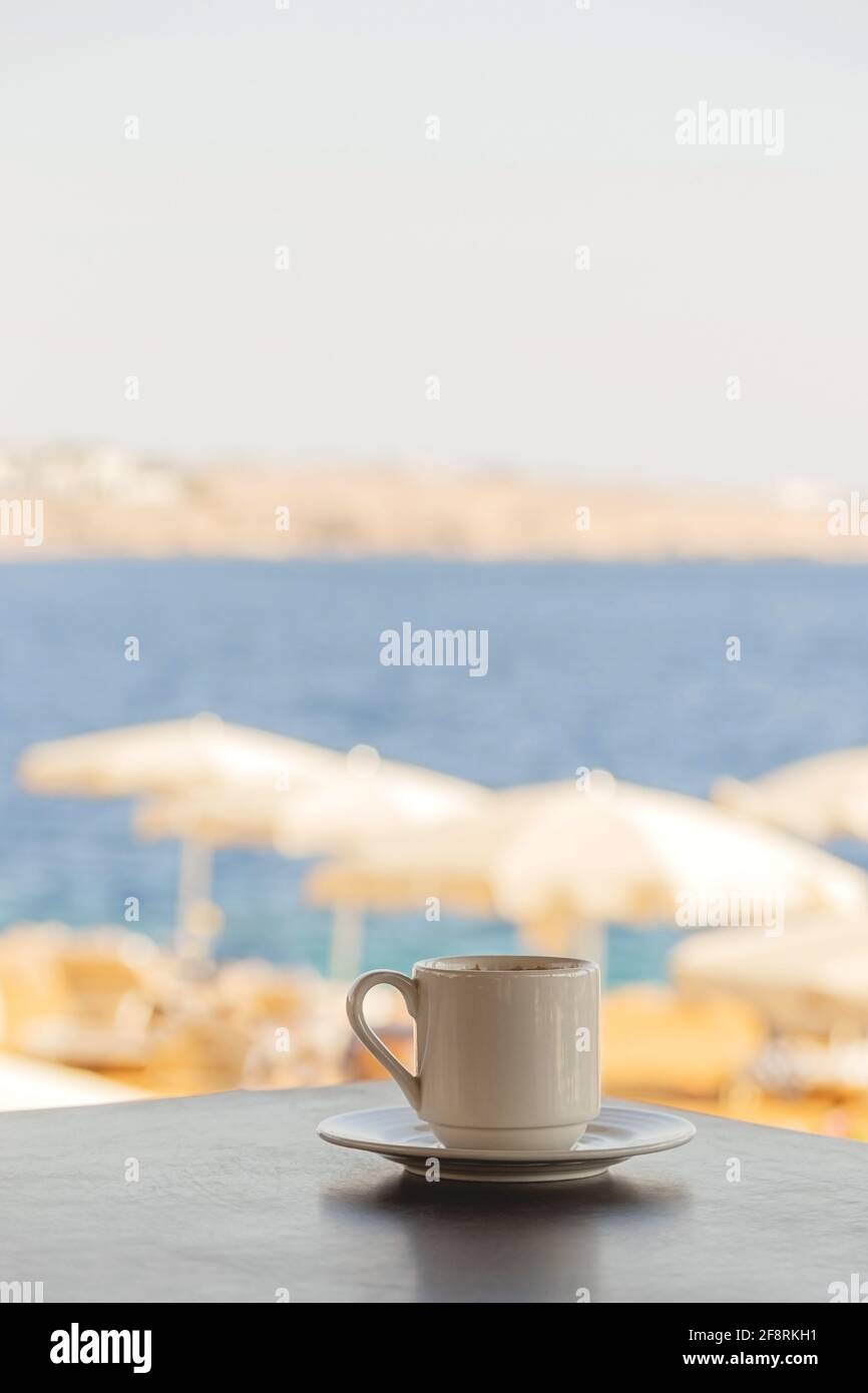 Une tasse de café blanc sur la table donnant sur la plage avec des parasols blancs et la mer bleue. Banque D'Images