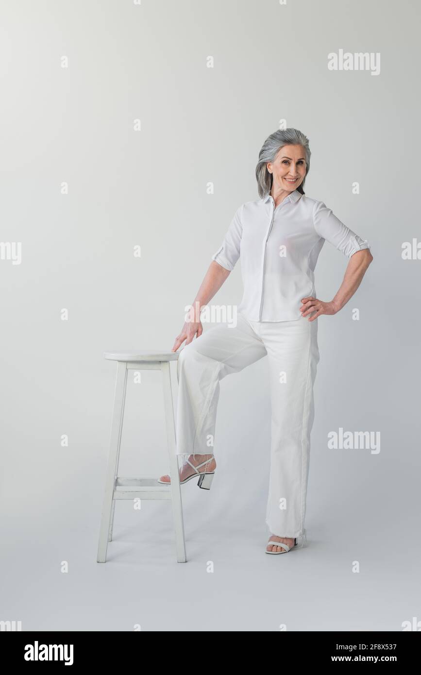 Femme mûre avec la main sur la hanche regardant l'appareil photo à proximité chaise sur fond gris Banque D'Images