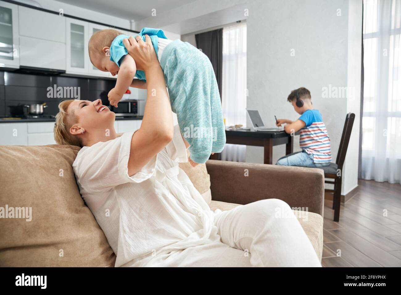 Une mère heureuse jouant avec son plus jeune fils sur un canapé tandis qu'une autre aînée est assise sur une table et utilise un ordinateur portable pour étudier. Femme avec deux fils séjournant à la maison. Banque D'Images