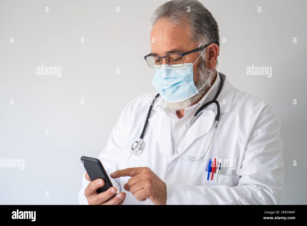 Portrait d'un médecin barbu en manteau blanc avec son smartphone sur fond blanc. Banque D'Images