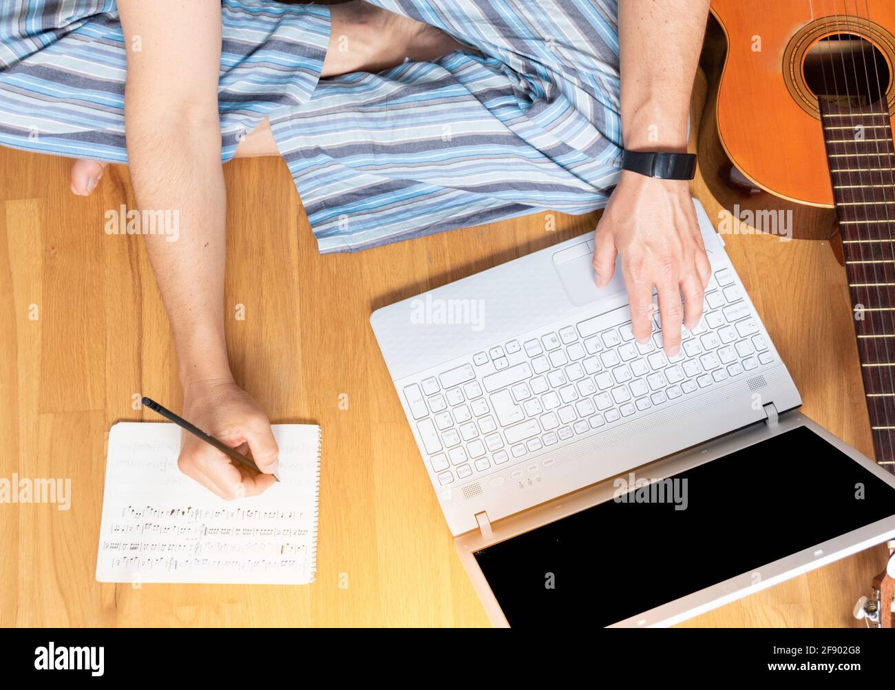 L'homme consulte son ordinateur portable et prend des notes. Il est assis sur le plancher en apprenant à jouer de la guitare. Banque D'Images