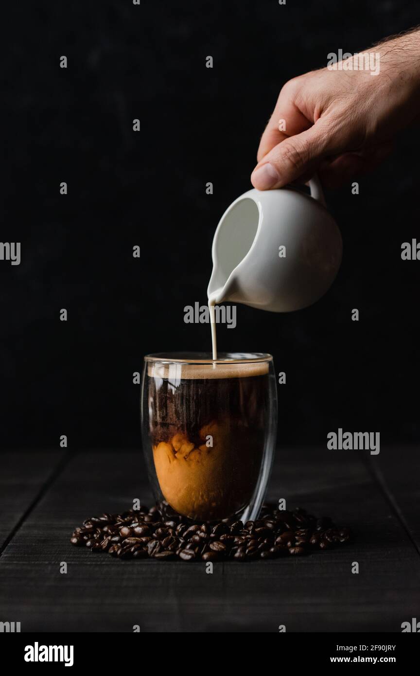 Versez la crème à la main dans une tasse de café en verre transparent sur fond noir. Banque D'Images