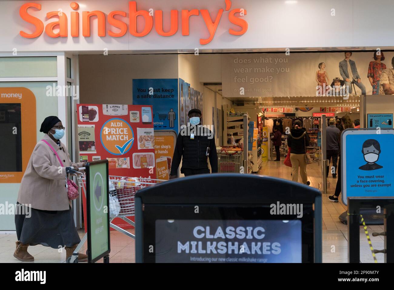 une casheuse pousse son chariot à l'entrée du magasin sainsbury, un agent de sécurité portant des supports de masque facial à l'avant du magasin Banque D'Images