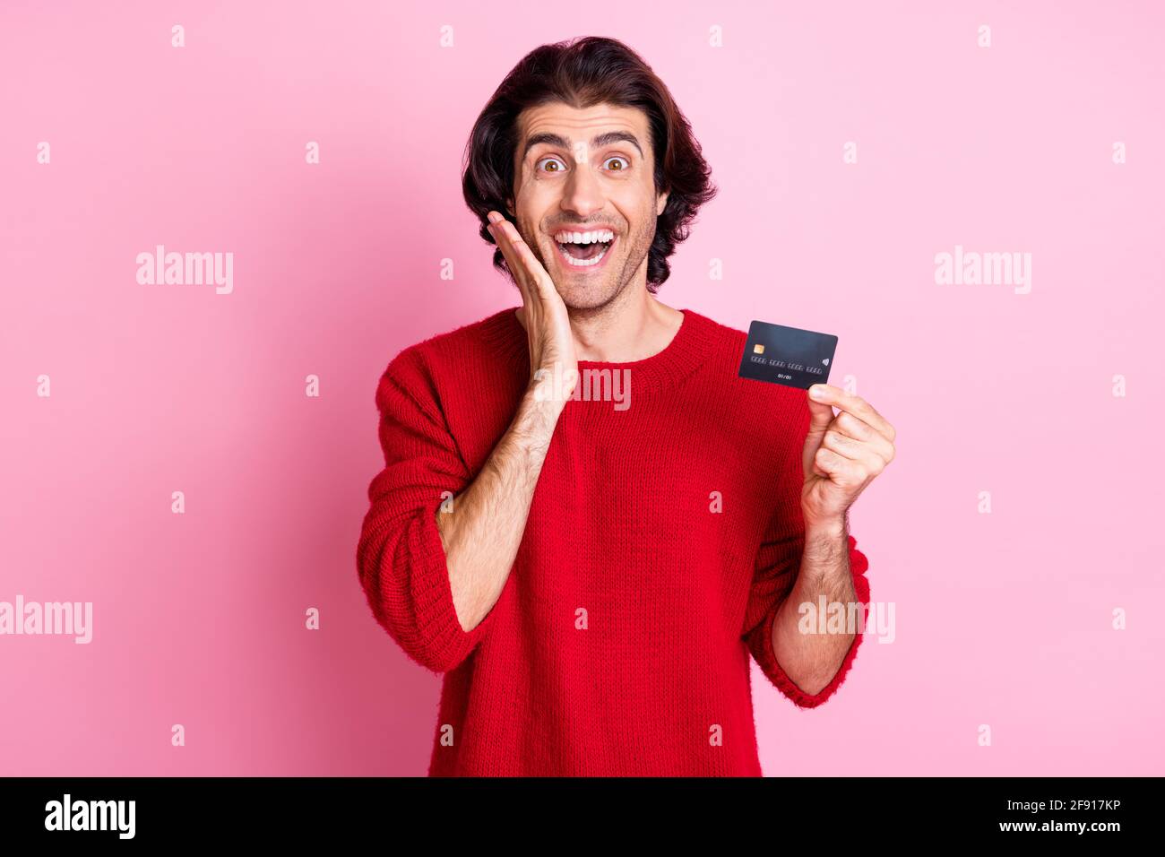 Photo portrait de l'homme choqué touchant le visage joue tenant crédit carte d'une main isolée sur fond rose pastel Banque D'Images