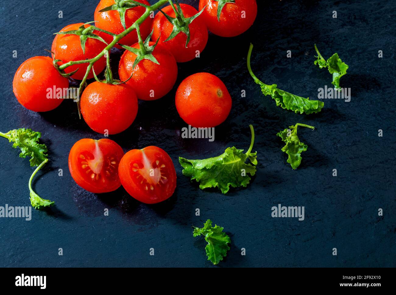 Feuilles vertes de choux de chou vert et tomates fraîches de vigne (cerise) avec des gouttes d'eau, brutes, entières et coupées en deux. Sur fond d'ardoise texturé noir. Ingredie Banque D'Images