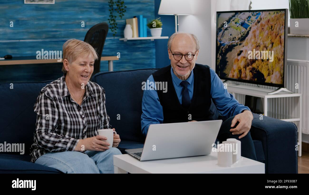 Un couple enthousiaste de retraités assis à la maison sur un canapé à l'aide d'un ordinateur portable parlant en vidéo avec des amis de la famille qui agite à la caméra pendant une réunion en ligne. Concept de style de vie de connexion en quarantaine Banque D'Images
