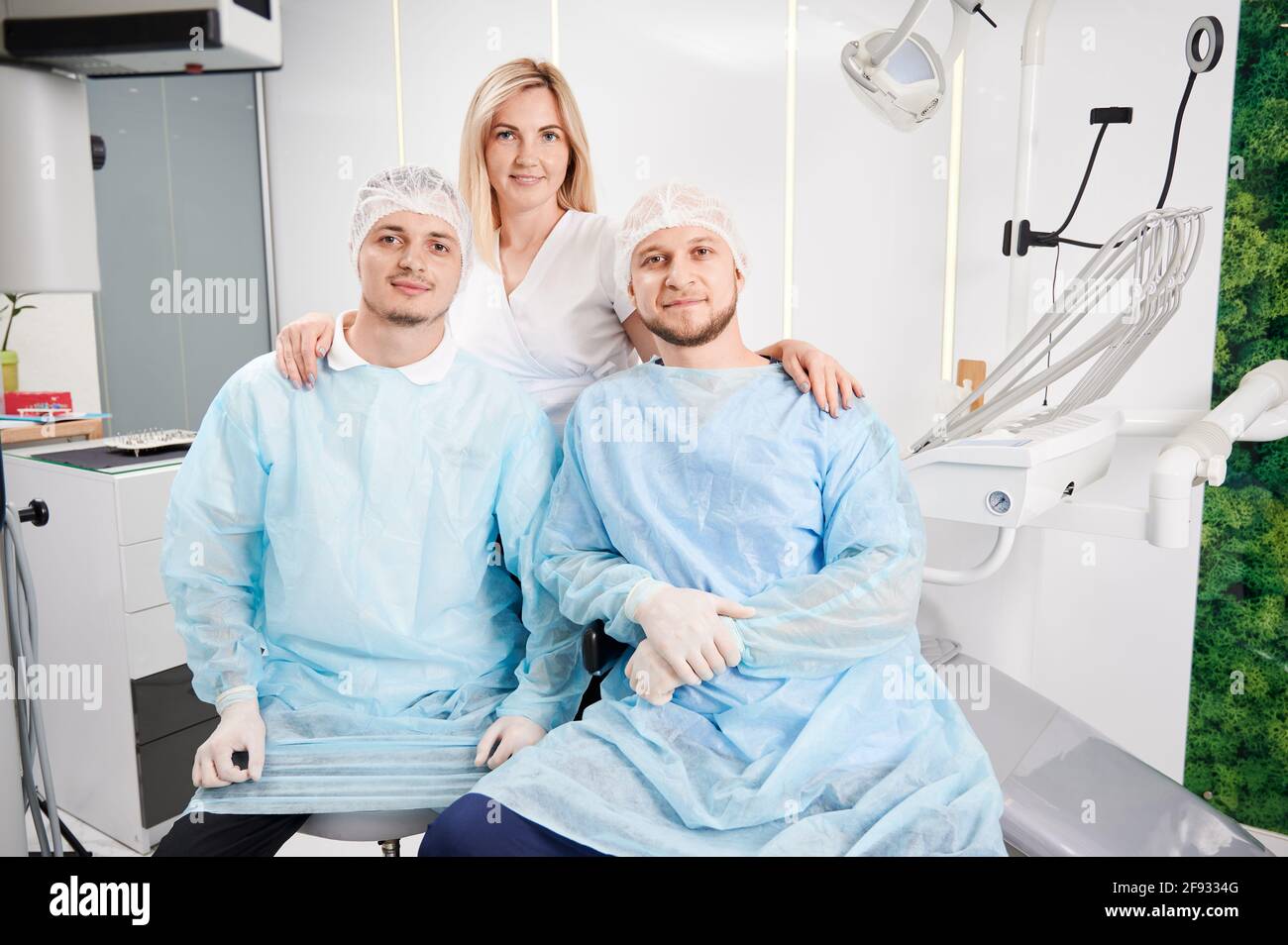 Vue de face des stomatologistes masculins dans des combinaisons de protection médicale regardant l'appareil photo et souriant tandis que charmante femme médecin debout derrière les hommes. Concept de la dentisterie et du personnel dentaire. Banque D'Images