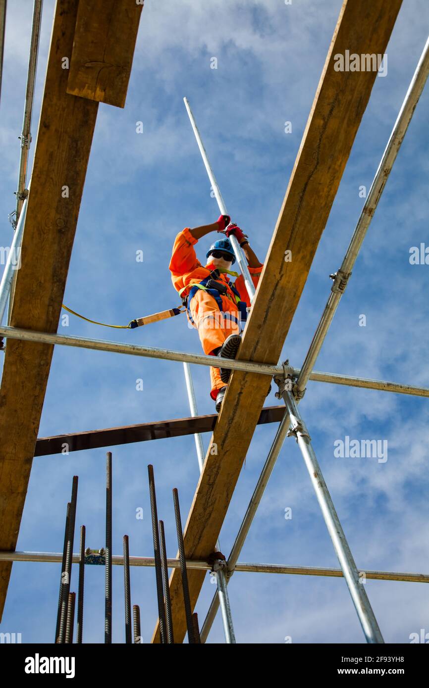 Grimpeur industriel dans des combinaisons orange et un casque de chantier vert assemblant des échafaudages par temps ensoleillé sur fond bleu ciel. Développement du conc. De renforcement Banque D'Images