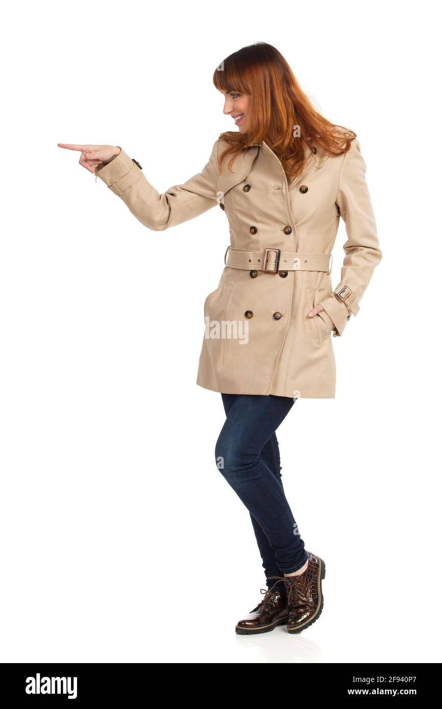 Jeune femme en beige manteau, jeans et chaussures brunes est debout, pointant ou poussant quelque chose. Vue latérale. Prise de vue en studio sur toute la longueur isolée sur blanc. Banque D'Images