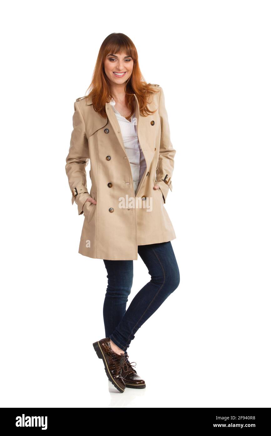 Jeune femme en beige manteau, jeans et chaussures marron est debout avec les mains dans les poches, souriant et regardant l'appareil photo. Vue avant. Prise de vue en studio Banque D'Images