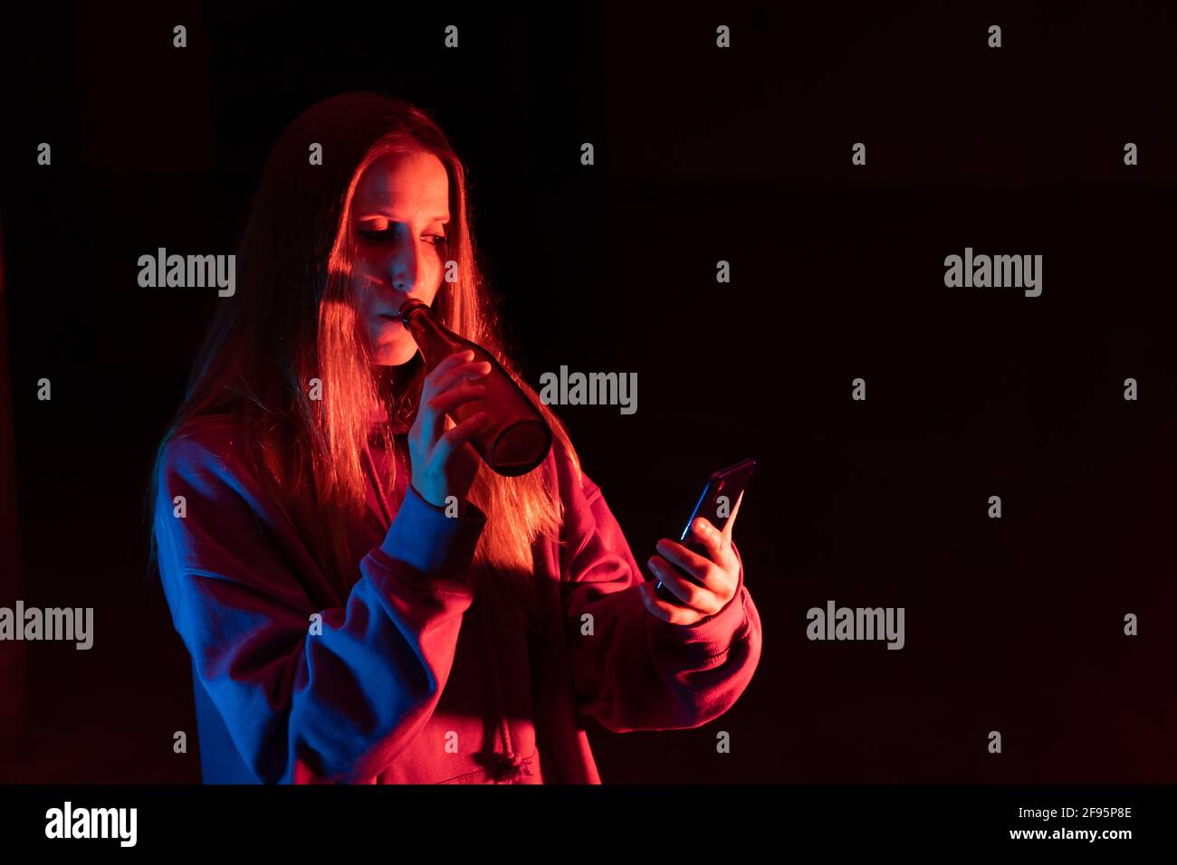 Une jeune fille boit une bière en regardant le smartphone Dans les mains.Portrait de la femme caucasienne sur le fond sombre du studio avec Lumières fluo colorées.nuit c Banque D'Images