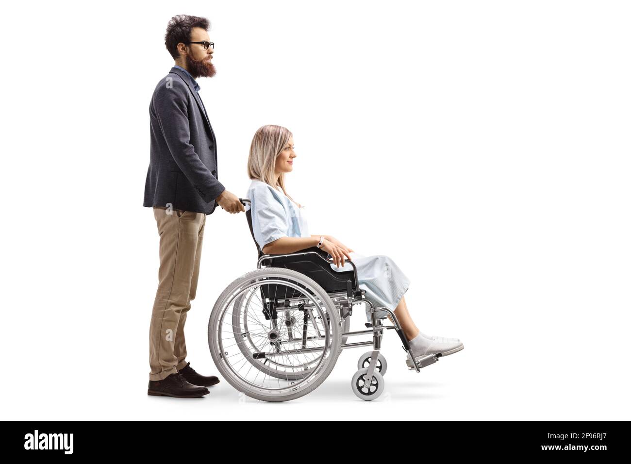 Prise de vue sur toute la longueur d'un homme barbu poussant un jeune femme patiente en fauteuil roulant isolée sur fond blanc Banque D'Images