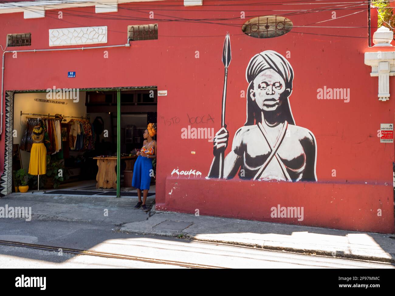 Photographie de rue: Jeune femme noire debout devant une boutique avec un graffiti sur le mur à côté d'elle d'une femme guerrier africaine qui ressemble à elle. Tourné avec Leica M10 à Rio de Janeiro Banque D'Images