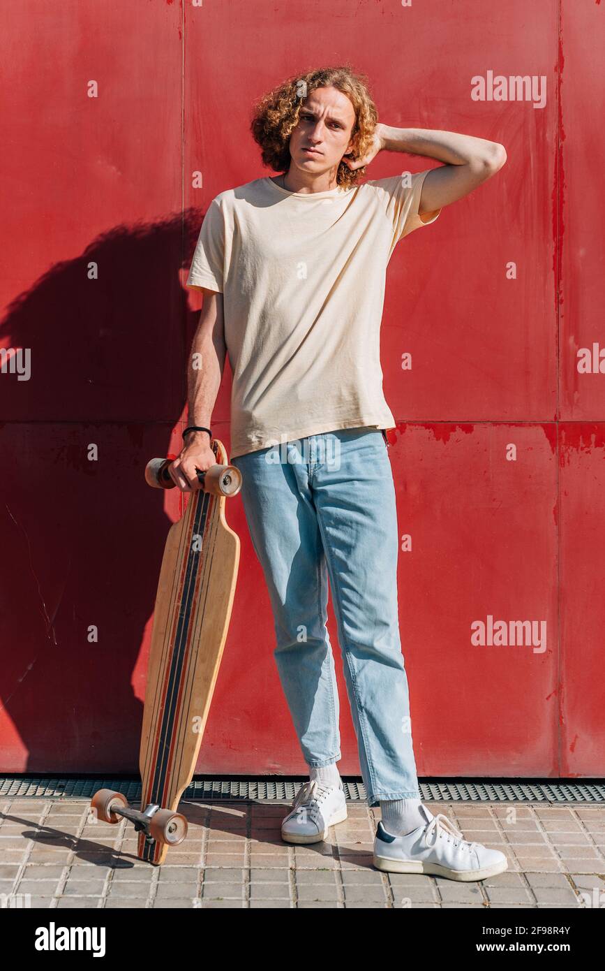 portrait vertical du corps d'un homme restant à prendre le skateboard avec la main. Il est jeune, avec une tête rouge et des robes décontractées. Arrière-plan rouge Banque D'Images