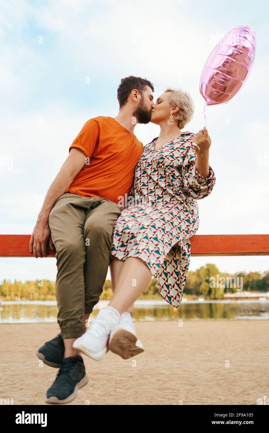 Homme et femme embrassant tout en étant assis et tenant un rose ballon en forme de coeur avec ciel sur fond Banque D'Images