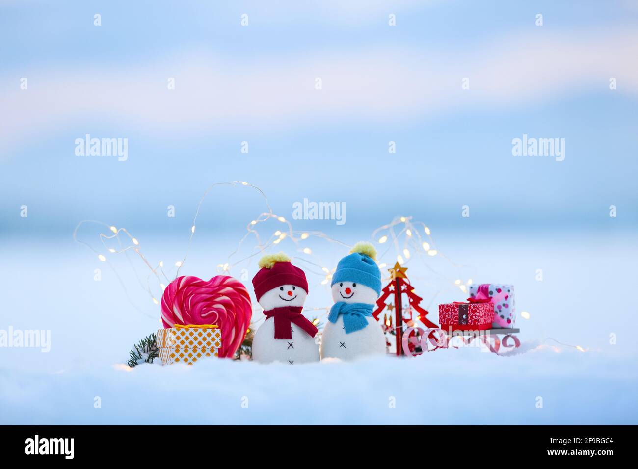 Amis bonhommes de neige restent avec un traîneau et une boîte cadeau sur la neige éclairée par des lumières de fées. Bonbons rouges à coeur blanc. Bonne Année. Composition de Noël. Banque D'Images