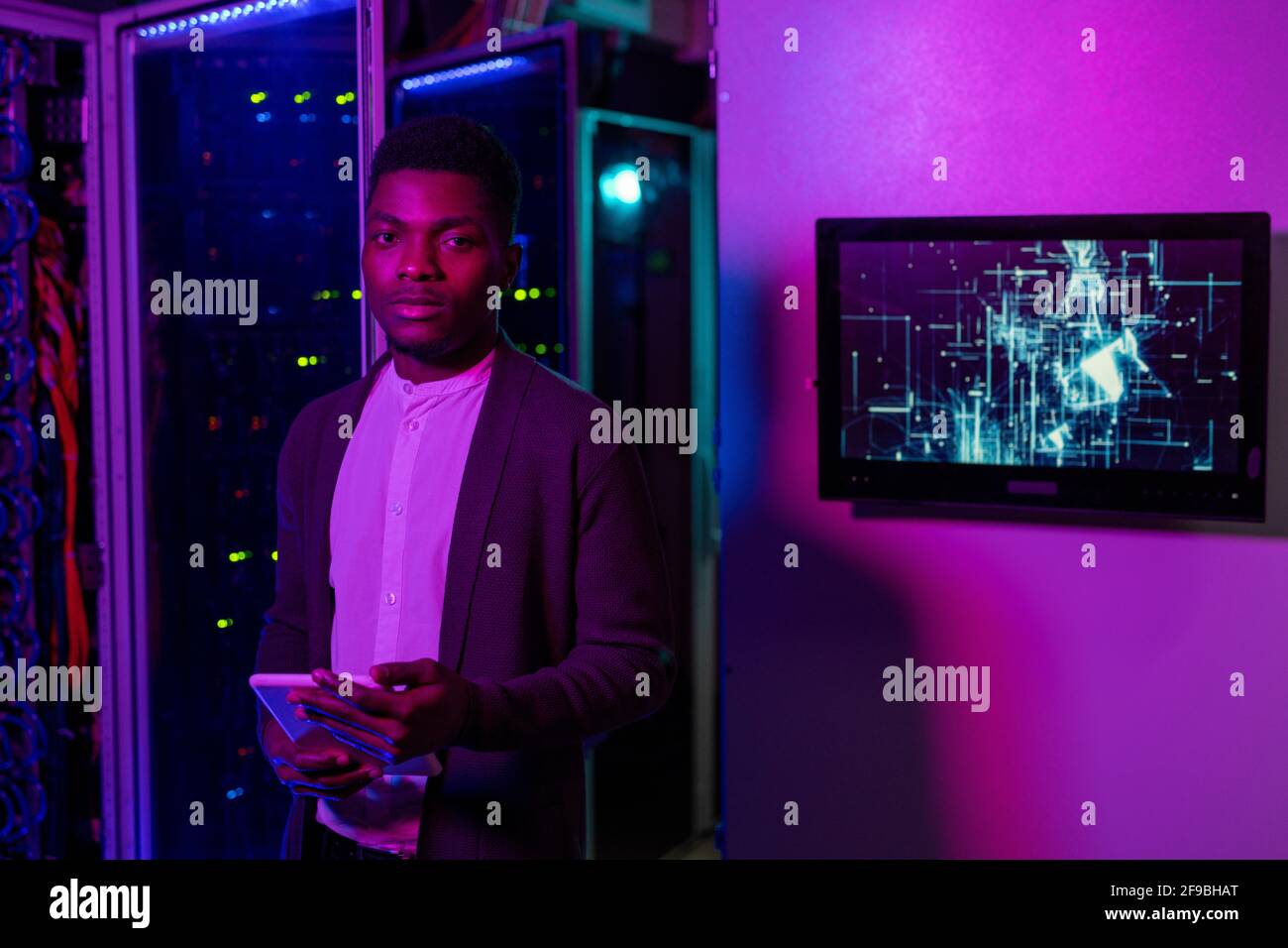 Portrait d'un jeune ingénieur INFORMATIQUE noir plein d'assurance dans un gilet debout avec tablette dans une pièce éclairée au néon foncé et travailler avec un superordinateur Banque D'Images