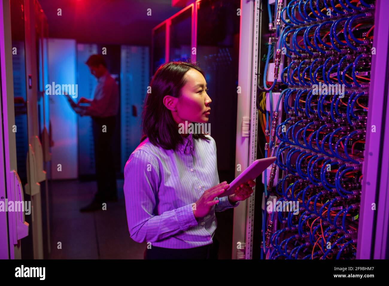 Jeune femme asiatique concentrée dans une chemise dépouillée debout au serveur armoire rack et utilisation de la tablette tout en maintenant le réseau dans le serveur chambre Banque D'Images
