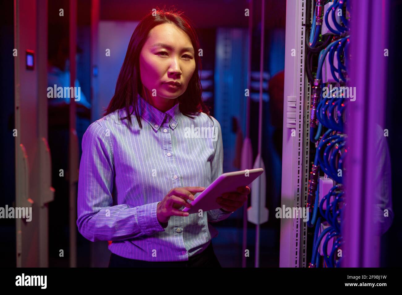 Portrait d'un ingénieur réseau asiatique sérieux qui effectue un dépannage tout en travaillant avec un superordinateur dans une salle de serveurs, lumière néon sombre Banque D'Images